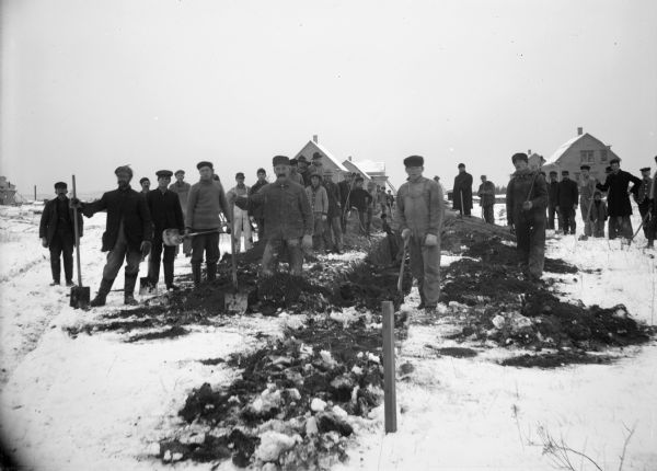 Outdoor view of a group of a large group of people gathered along a trench. Snow is on the ground, and buildings are in the background. Several of the men are holding shovels. Envelope labeled "Vandrieul."