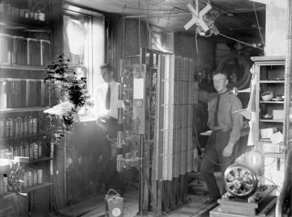 Interior view of a room with telephone equipment and two men standing, one man by a window on the left, and another man near equipment on the right.