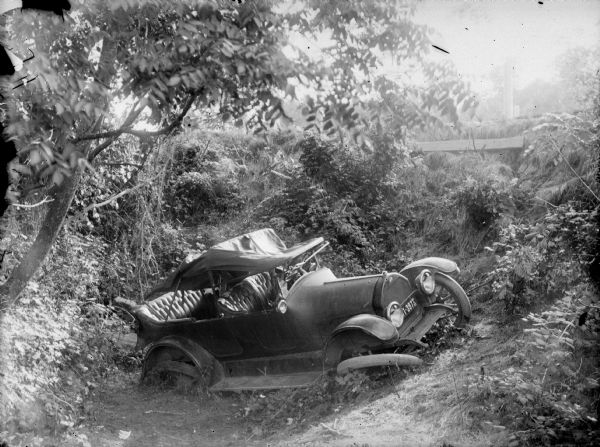 View looking down towards of an automobile resting in a roadside ditch. The soft roof cover is collapsed, and the wheels on the passenger side are bent. The Wisconsin license plate has a date of 1922.
