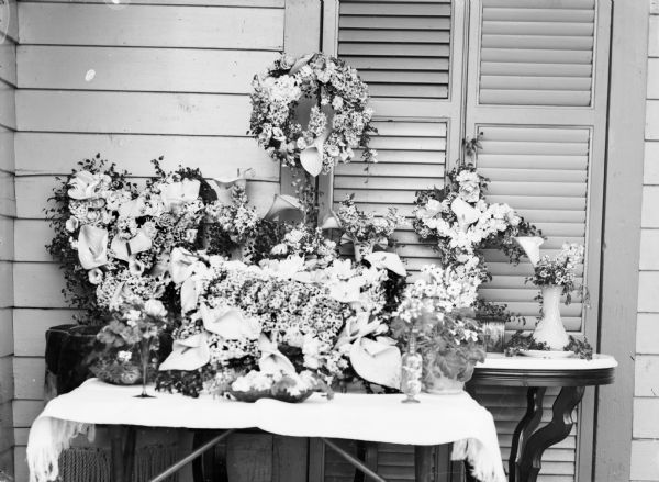 View of funeral floral arrangements set up on tables next to a house with shutters.