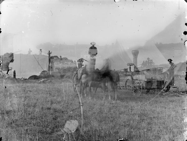 In the foreground is a farming scene with a group of men in the background on the left, a man standing on a cart with two horses, and another man on the right is standing on a steam-powered tractor. The other scene in the background has a tent, and buildings in the background.