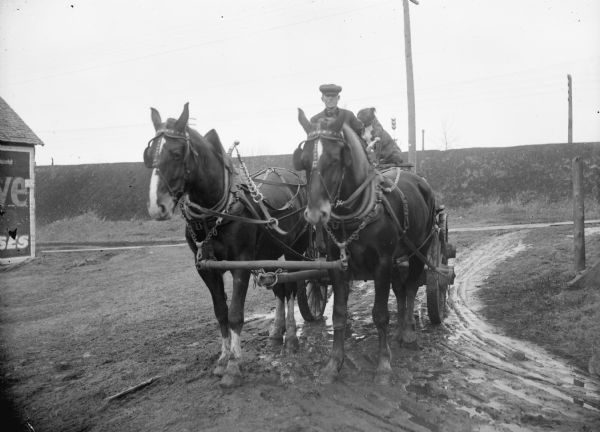 A man and his dog are sitting on a cart pulled by two horses at a rural intersection on a muddy road. There is a corner of a building with a billboard on the side of it on the far left. In the background is the steep bank of a railroad, with a railroad signal.