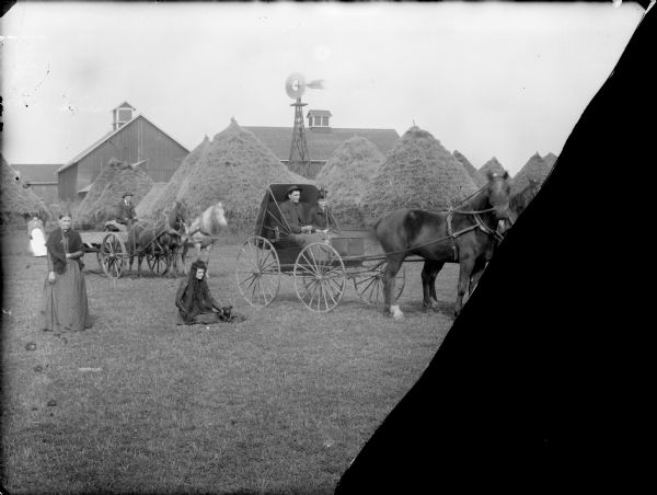 A farm scene with a group of people posing in front of farm buildings, tall, cone-shaped haystacks, and a windmill. In the center is a man and woman sitting in a horse-drawn buggy, a man sitting in a wagon pulled by two horses. A woman is standing on the left, and another woman is standing in the background behind her. A woman is sitting on the grass in the center with a dog beside her.