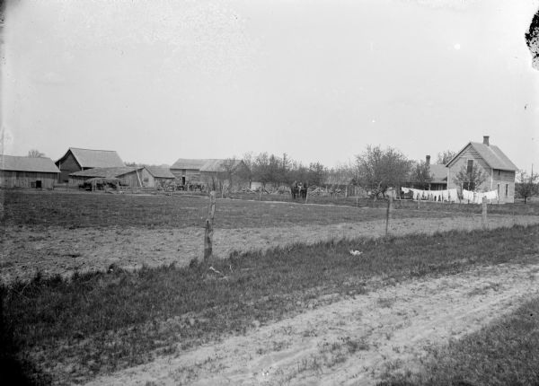 View across road and field towards a farm. The farmhouse is on the right, with farm buildings on the left. A two-horse cart is in the center.