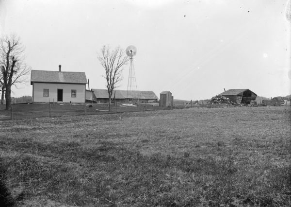 A view of a farm with a house on the left, a windmill in the center, and a dilapidated building on the right.