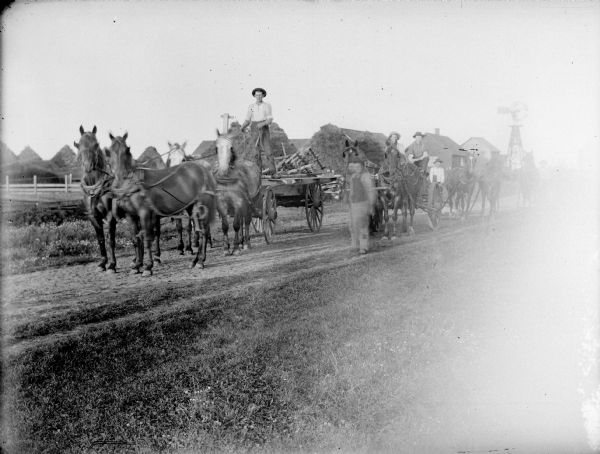 View from side of road towards men with horses and carts. A farm with large, cone-shaped haystacks and a windmill is in the background.