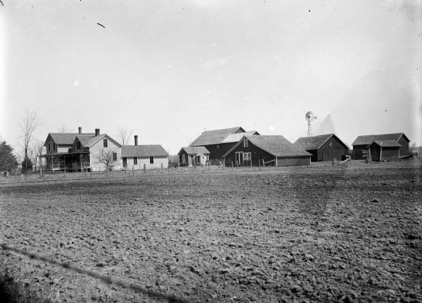 View of a farm from across a field of dirt. The house is on the left, with several farm buildings and a windmill on the right.