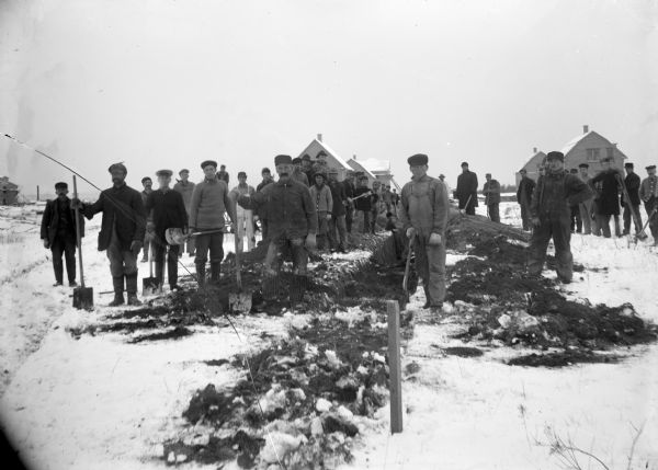 Outdoor group portrait of a large group of men standing in the snow, many of them holding shovels. A ditch has been dug in the center. Houses are in the background.