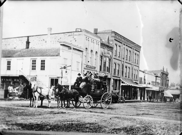 Copy photograph of a crowded stagecoach is being pulled by four horses through the intersection of First and Main Streets.