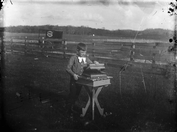 View towards a boy standing outdoors in the grass leaning against a table with photo albums stacked on it. A fence is in the background, and a buggy is parked on the road behind it. There is a sign (unreadable) painted on the side of the buggy.