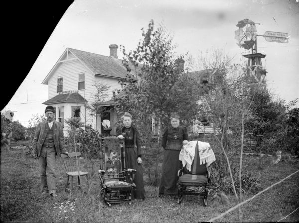 Group portrait of a man and two women, each one posing standing next to a chair in the front yard of a home. A windmill is on the right with a sign that reads: "Halladay Standard U.S. Wind Eng. & Pump Co., Batavia, Ills." Behind the group, on the porch, is another chair.