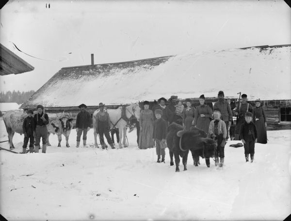 Outdoor group portrait of people standing in the snow with horses and cattle. A long log building is behind them.