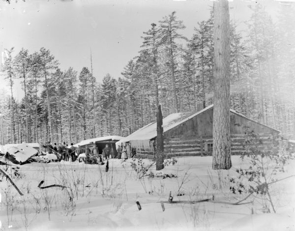 View across snow towards a group of loggers, including two women, one holding a child in her arms, at a logging camp in winter.