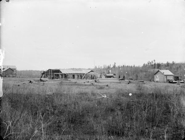 View across field of scrub grass towards a group of men, some in work clothes and other men wearing suits, standing in a lumberyard at a logging camp. There is a bell on a post near the building on the right, and two wagons are parked nearby. A poster on the wall of the building is for "A.F. Werner, Leading Clothier Hatter Furnisher."