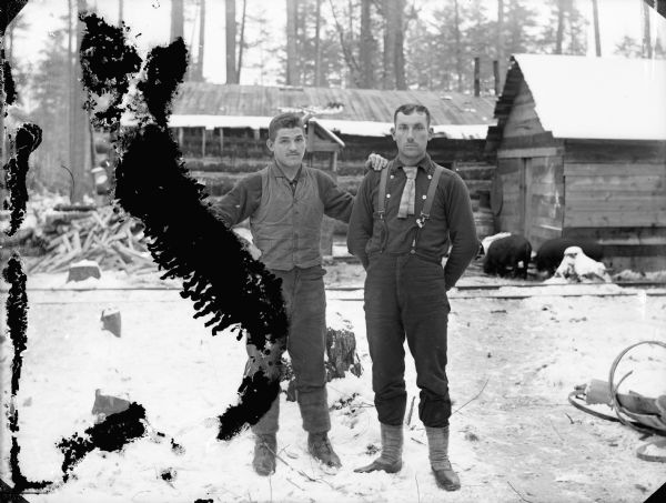 Two men are standing in the snow at a logging camp. In the background are railroad tracks, and two pigs foraging around buildings.