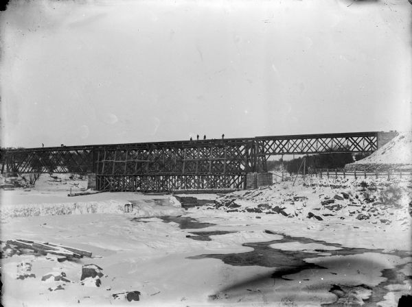 View across a frozen river towards a railroad bridge. A group of men are standing on top of the bridge in the center, and two other men are on the left.