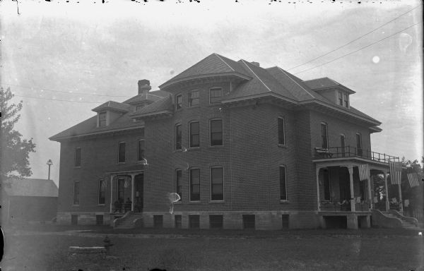 Exterior view of the Jackson County Home. Flags are hanging from the front porch, which has a balcony above. Men are sitting on the side landing on the left, which also has flags mounted on the porch columns. There is a bell on a post behind a small building in the backyard on the left.