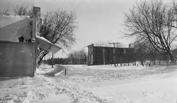 Winter scene looking east on Main Street (Highway 42), with wooden commercial buildings on both sides of the street. The building on the left, 4168 Main Street, has a large wooden awning and a snow drift in front. It housed general stores under the Lundberg, Vorous, Krause and Alwes names. The building in the background, now 4153 Main Street, housed the Levi Vorous store in front and Noble's blacksmith shop in the rear.