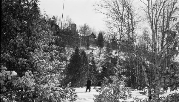 Ferdinand Hotz poses on snowshoes at the base of a bluff. The Hotz cottage complex, built in 1912-3, is seen atop the bluff.  The complex included three cottages, a log outhouse, pumphouse, and stone garage with tower. There are snow-covered cedars in the foreground.