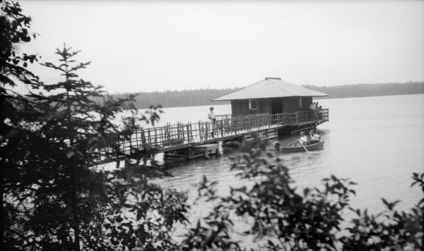 View from shoreline of a boy rowing a small boat with a girl seated aft as a woman watches them from the walkway to a boathouse. The walkway, with its rustic railing, extends around the boathouse. Other people are gathered at the corner of the boathouse.