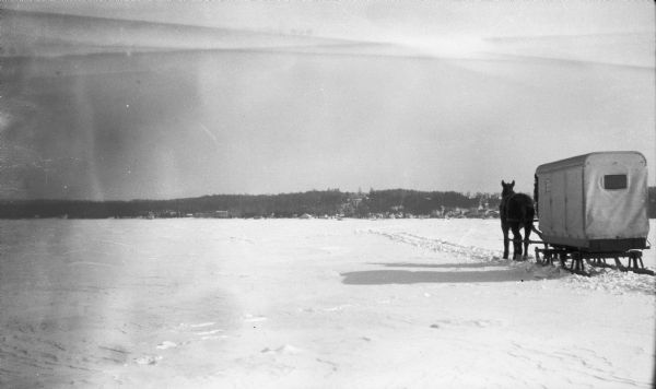 A single horse pulls an enclosed sleigh across a frozen Door County harbor. Houses and a church are visible onshore.