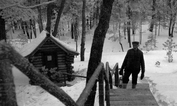 The caretaker, carrying a lantern, poses on stairs leading up from the log outhouse at the Hotz Europe Lake compound. There is hoar frost on the trees and stair railing.