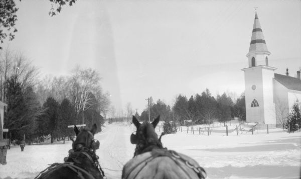 View from behind of two horses (probably pulling a sleigh) on a snowy road, now Highway 42, near Zion Lutheran Church in Sister Bay. The horse on the left wears sleigh bells. A sign on the road reads: BIRCHWOO(?) HALL RESORT."