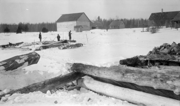 Three men pose on logs as a fourth walks toward them; there are axes and snowshoes on the ground. A large log barn, outhouse, and other buildings are in the background.