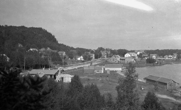 Elevated view of Fish Creek from the east; the bluff rises on the left, the harbor is on the right. The cottage in the foreground with pergola and lattice is the photographer's first Fish Creek Cottage. The two paired buildings on the waterfront were bathhouses and boathouses for the Thorp and Welcker resorts. The Welcker Casino is the largest building, with a dark gable, in the background.