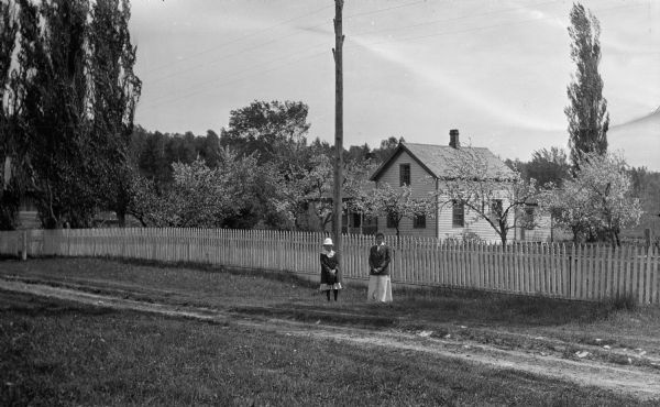 Alice Hotz Apfelbach, right, and her younger sister Margaret Hotz pose in front of a picket fence along a dirt lane which is now Highway 42.  Behind the fence is a wood frame farmhouse with fruit trees blooming in the yard. The house, part of a farmstead originally owned by Levi Vorous, was given to Alice and her husband George as a wedding present in 1923.  The house has been moved and the Fish Creek Condominiums stand on the property.  The tall tree to the left of the utility pole was an elm which served as a landmark for many years.