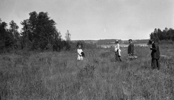 A couple, center, and two girls pose in an opening on high ground. The girl on the left is the photographer's daughter, Margaret. The man carries two picnic baskets. A bay and village are visible in the distance.