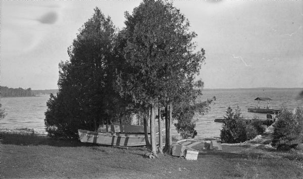 View from lawn of small boats which have been pulled onto the shore alongside a pier at North Bay. White cedars (arbor vitae) grow along the pier and on shore.