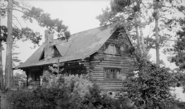 View of the rear of the Hotz cottage, showing a stone chimney which divides a dormer. There are large pine trees around the cottage.