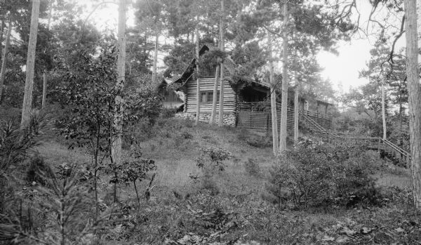 Three-quarter view of the Hotz cottage showing a side and the large porch which faced Europe Lake. The stone foundation is visible on the side; the porch has a rustic railing and lattice work. There is a log sided structure in the background behind the cottage.