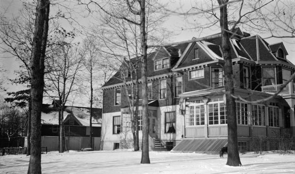 Three-quarter view of the Hotz home at 1072 Sheridan Road in winter. There is a large barn behind the house; a dog is partially visible behind a tree.