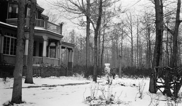 A girl with a large bow in her hair stands beside a snowman in the front yard of the Hotz residence at 1072 Sheridan Road. The house has a large front porch and there is a rustic fence on the right.