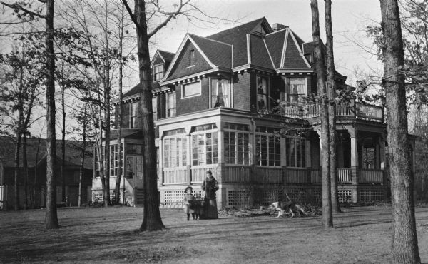 Mrs. Ferdinand (Clothilde) Hotz poses with two of her daughters in front of their house. A dog walks toward them from the right. The large shingle-style house has multiple porches, gables and dormers, with dentil molding under the eaves. There is a large barn in the background.