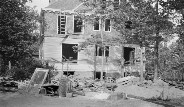 Lumber, wheelbarrows, a screen for sifting sand, and portions of scaffolding clutter the area around a new addition to the rear of the Hotz residence. There is narrow siding on the first story, with shingles on the second story and gable.  The addition included a billiard room (left) and music room (right), as well as sleeping quarters for servants on the second floor.