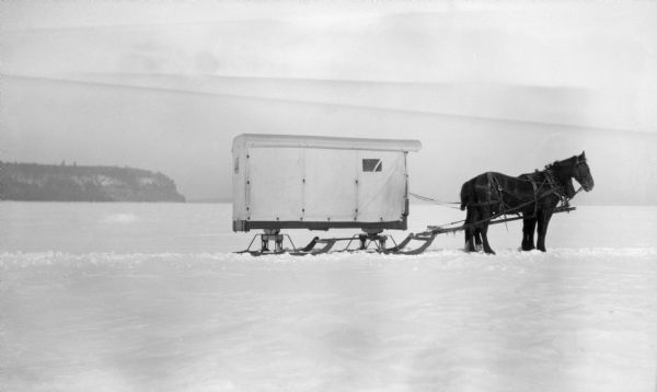 Winter scene with a team of horses standing harnessed to an enclosed bobsled on snow- and ice-covered Ephraim Bay.