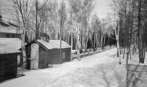 A small log sided building with roof ventilator, among other buildings, stands near the Wilms house along Cottage Row Road outside of Fish Creek. A low stone wall, birch and evergreen trees line the road.