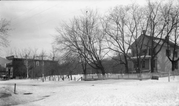 View looking southeast at the corner of Main (Egg Harbor Road) and Spruce Streets. Snow is on the ground. A neoclassical house (the Noble house) with front porch is standing among mature trees behind a picket fence; a two-story wood frame commercial building is on the left, with a "Garage" sign in front.