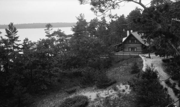Elevated view of the log Hotz cottage on Europe Lake, seen in the background. The photographer climbed a tree to capture this view. A dirt road leads to the cottage, which has a rustic railing and latticework.