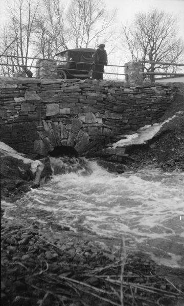 The photographer's wife Clothilde leans on a railing, looking at water rushing through a stone culvert on Fish Creek. Their car is parked on the roadway. A young boy or girl is standing on the other side of the bridge.