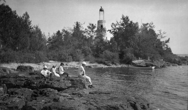 Ferdinand Leonard (Fedy) Hotz, far left, sisters Helen, left, and Alice, sit with Thomas Toft on the rocky shore near the Baileys Harbor lighthouse. A rowboat is moored in the background.