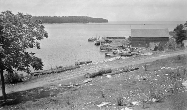 View from hill looking down at a wood framed warehouse building on a pier with boats docked alongside. Near the warehouse are fish net drying reels. Wooded bluffs are on the opposite shore of the bay. Three girls or women stand near a large log at the water's edge. A narrow road follows the shoreline.