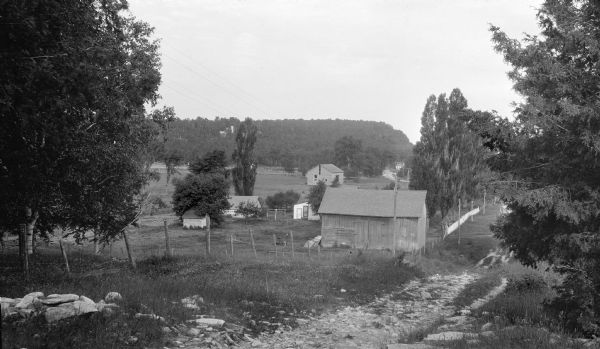 The barn and other outbuildings of the Vorous/Apfelbach farm stand along a rocky lane, now Highway 42.  There is a row of poplar trees along a picket fence in front of the house, which is mostly hidden by the barn. The stone tower and buildings of the Hotz compound are visible on the bluff beyond.