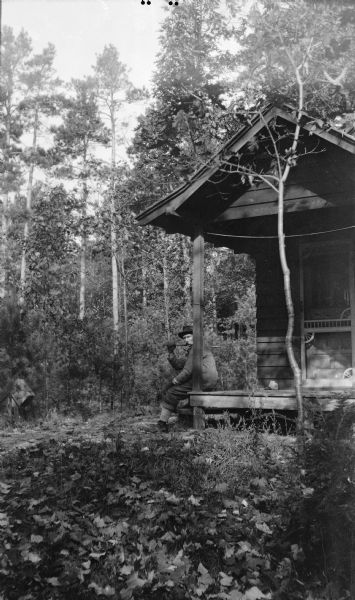 David Kincaid sits on the front porch of his rustic cabin in the woods, at the entrance gate to "Hotz Trail" Road near Europe Lake. He holds an axe over his shoulder.