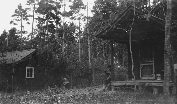David Kincaid sits on the porch of a rustic cabin in the woods, at the entrance gate of "Hotz Trail" Road near Europe Lake. He holds an axe over his shoulder.