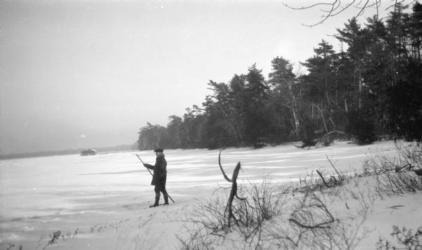 A man with a walking stick stands on the shore of frozen Europe Lake. The Hotz boat house and pier are in the background.