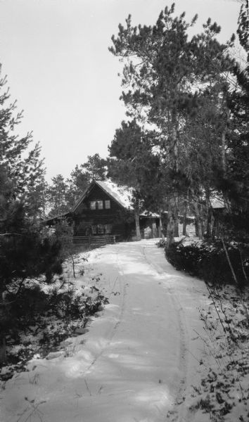 A man stands on the rustic porch of the Hotz cottage on Europe Lake. There is a single set of tire tracks in the snow on the drive.